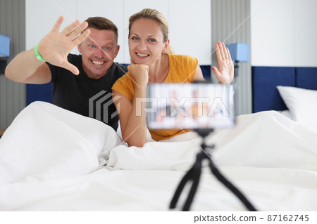 Couple in bed waving at smartphone camera on tripod in hotel Couple in bed waving at smartphone camera on tripod in hotel 87162745