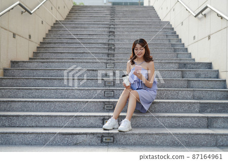 Cheerful young Asian woman holding takeaway coffee cup, using mobile phone 87164961