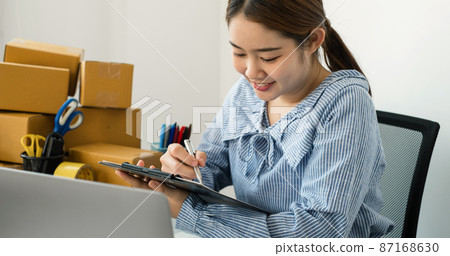 Young Asian woman business owner with many parcel boxes on the table happy online sales job, use the laptop, get an order from customers, take notes, and make arrangements for delivery by post 87168630