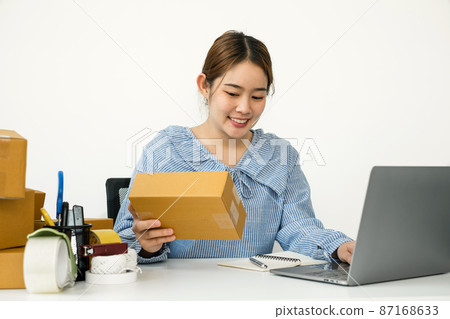 Young Asian woman business owner with many parcel boxes on the table happy online sales job, use the laptop, get an order from customers, take notes, and make arrangements for delivery by post 87168633