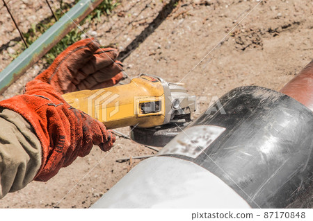 The hands of an industrial worker in working mittens use an electric-grinding equipment tool on a polyethylene water pipe on a construction site The hands of an industrial worker in working mittens use an electric-grinding equipment tool on a polyethylene water pipe on a construction site 87170848