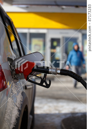 silver metallic color car refueling on winter day gas station - close-up with selective focus and blurry man silhouette in the background 87173283