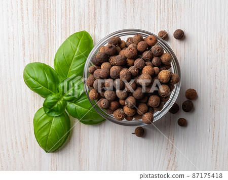 Glass bowl full of allspice jamaica pepper near fresh basil leaves on a white wooden table. Glass bowl full of allspice jamaica pepper near fresh basil leaves on a white wooden table. 87175418
