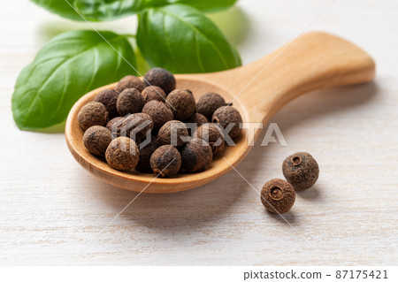 Allspice jamaica pepper in a wooden spoon near fresh basil leaves on a white wooden table. 87175421