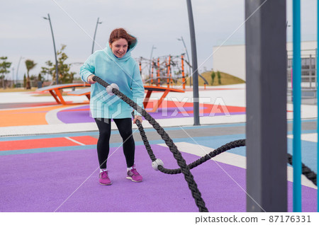 Caucasian woman in a mint sweatshirt is training with battle ropes at the sports ground. Caucasian woman in a mint sweatshirt is training with battle ropes at the sports ground. 87176331