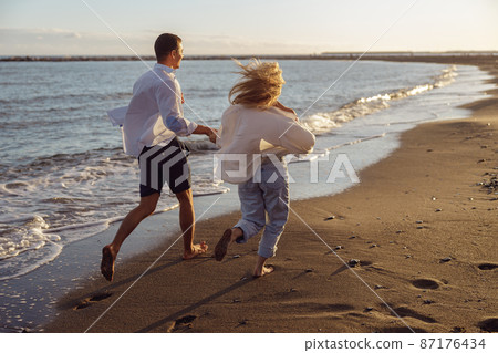 Back photo of man and woman running holding hands along the ocean Back photo of man and woman running holding hands along the ocean 87176434