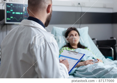 Doctor holding clipboard with latest test results after surgery talking with patient recieving oxygen Doctor holding clipboard with latest test results after surgery talking with patient recieving oxygen 87177000