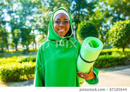 latin black woman in green hijab with bright make up and piercing nose holg yoga mat outdoor in summer park 87185644