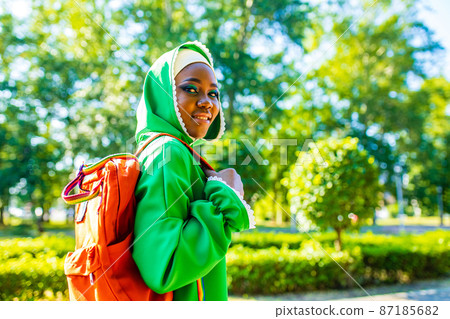 modern afro woman in green hijab with bright make up and piercing nose holg orabge backpack in park 87185682