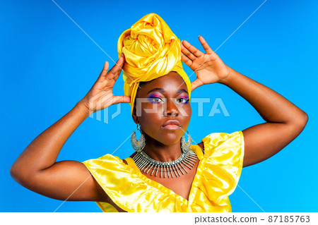 beautiful african woman in yellow silk turban on head and dress with silver neckless and earrings , nose piercing in blue studio background beautiful african woman in yellow silk turban on head and dress with silver neckless and earrings , nose piercing in blue studio background 87185763