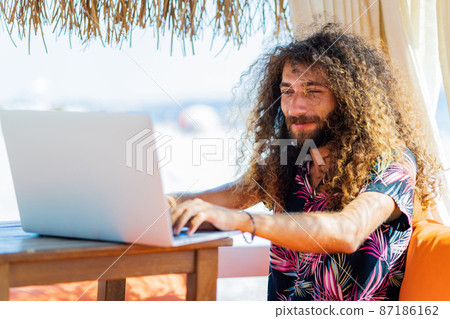 long curly haired man sitting in beach and working laptop vacation long curly haired man sitting in beach and working laptop vacation 87186162