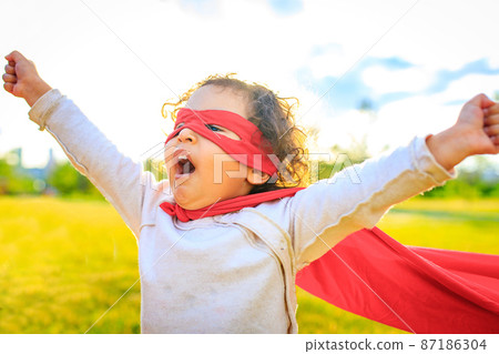 african american little girl in red costume and eyes mask playing a hero in summer park 87186304