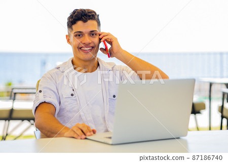 man in white t-shirt talking by phone while remote working at sunny beach in cafe man in white t-shirt talking by phone while remote working at sunny beach in cafe 87186740