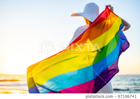 woman with straw white hat holding a gay pride rainbow flag blowing in the wind on beach with golden sunset woman with straw white hat holding a gay pride rainbow flag blowing in the wind on beach with golden sunset 87186741