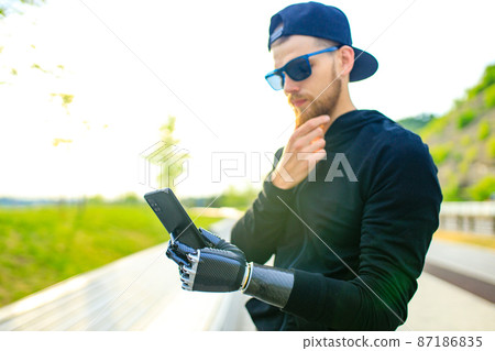 young bearded man with metal arm prosthetic outdoor looking at screen of smartphone young bearded man with metal arm prosthetic outdoor looking at screen of smartphone 87186835
