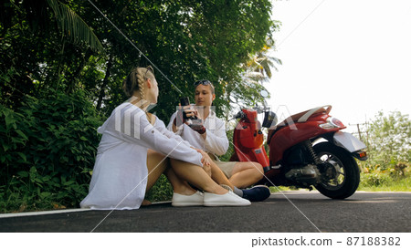 Two caucasian tourist woman man sit near red scooter. Make it selfie. Love couple on motorbike in white clothes on forest road trail trip. Two caucasian tourist woman man sit near red scooter. Make it selfie. Love couple on motorbike in white clothes on forest road trail trip. 87188382