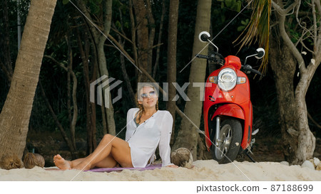 Scooter road trip. Woman alone on red motorbike in white clothes on sand beach by ocean. One girl caucasian tourist walk near tropical palm tree. Scooter road trip. Woman alone on red motorbike in white clothes on sand beach by ocean. One girl caucasian tourist walk near tropical palm tree. 87188699