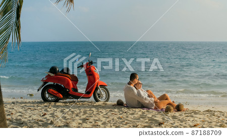 Scooter road trip. Lovely couple on red motorbike in white clothes on sand beach. People walking near the tropical palm trees, sea. Motorcycle rent. 87188709