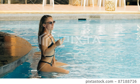 The pretty woman drinking blue cocktail alcohol liquor and sit at the bar. Cocktail bar in the swimming pool. Portrait of sexy girl in pool outdoor. 87188716