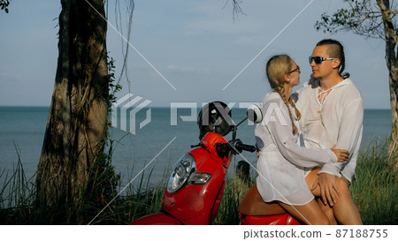 Love couple on red motorbike in white clothes on forest road trail trip. Two caucasian tourist woman and man kiss hugs sit on scooter. 87188755