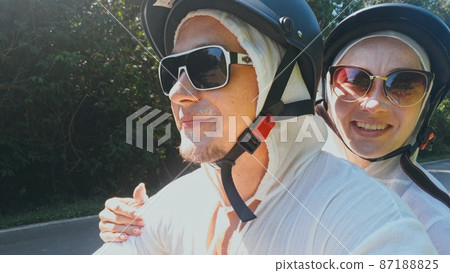 Two caucasian tourist woman man drive on red scooter. Make it selfie. Love couple on motorbike in white clothes to go on forest road trail trip. Two caucasian tourist woman man drive on red scooter. Make it selfie. Love couple on motorbike in white clothes to go on forest road trail trip. 87188825