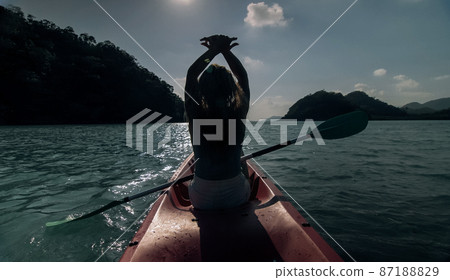 Silhouette of woman tourist raising hands while sailing in canoe along sea bay water to distant hilly island backside view. Silhouette of woman tourist raising hands while sailing in canoe along sea bay water to distant hilly island backside view. 87188829