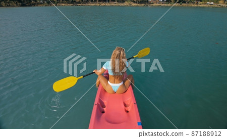 Seductive blonde woman in blue swimsuit sails on pink plastic kayak along azure sea at exotic resort backside upper above view. Seductive blonde woman in blue swimsuit sails on pink plastic kayak along azure sea at exotic resort backside upper above view. 87188912
