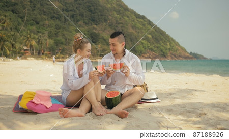 The cheerful love couple holding and eating slices of watermelon on tropical sand beach sea. Romantic lovers two people spend summer weekend. The cheerful love couple holding and eating slices of watermelon on tropical sand beach sea. Romantic lovers two people spend summer weekend. 87188926