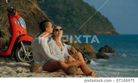 Scooter road trip. Lovely couple on red motorbike in white clothes on sand beach. People walking near the tropical palm trees, sea. Motorcycle rent. 87188975