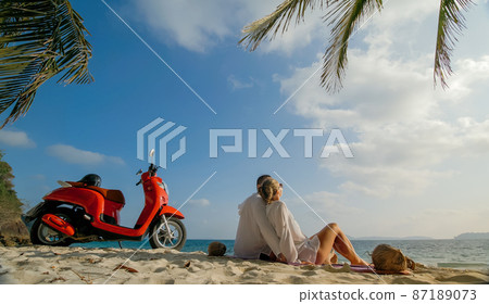 Scooter road trip. Lovely couple on red motorbike in white clothes on sand beach. People walking near the tropical palm trees, sea. Motorcycle rent. 87189073