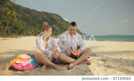 The cheerful love couple holding and eating slices of watermelon on tropical sand beach sea. Romantic lovers two people spend summer weekend. 87189088
