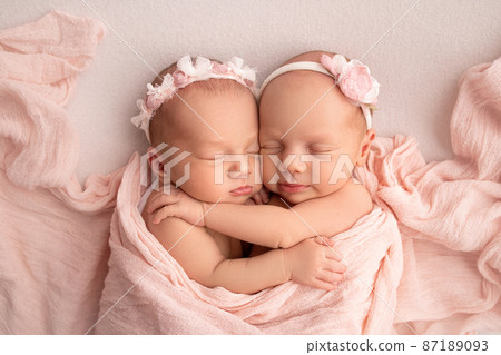 Tiny newborn twin girls. A newborn twin sleeps next to his sister. Newborn twin girls on the background of a pink blanket with pink bandages. The girls gently hug and kiss their sister in a cute pose. Tiny newborn twin girls. A newborn twin sleeps next to his sister. Newborn twin girls on the background of a pink blanket with pink bandages. The girls gently hug and kiss their sister in a cute pose. 87189093