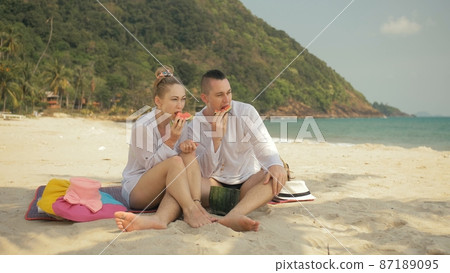 The cheerful love couple holding and eating slices of watermelon on tropical sand beach sea. Romantic lovers two people spend summer weekend. The cheerful love couple holding and eating slices of watermelon on tropical sand beach sea. Romantic lovers two people spend summer weekend. 87189095