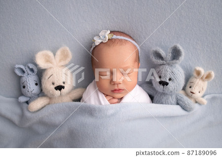 Sleeping newborn girl in the first days of life in a white cocoon with a white bandage and a blue flower on a blue background. Studio macro photography, portrait of a newborn ballerina, toys bunnies. Sleeping newborn girl in the first days of life in a white cocoon with a white bandage and a blue flower on a blue background. Studio macro photography, portrait of a newborn ballerina, toys bunnies. 87189096