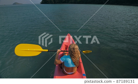 Seductive blonde woman in blue swimsuit sails on pink plastic kayak along azure sea at exotic resort backside upper above view. 87189131