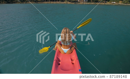 Seductive blonde woman in blue swimsuit sails on pink plastic kayak along azure sea at exotic resort backside upper above view. 87189148