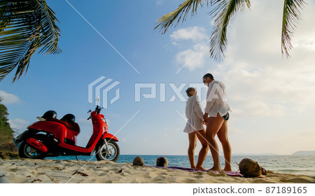 Scooter road trip. Lovely couple on red motorbike in white clothes on sand beach. People walking near the tropical palm trees, sea. Motorcycle rent. Scooter road trip. Lovely couple on red motorbike in white clothes on sand beach. People walking near the tropical palm trees, sea. Motorcycle rent. 87189165