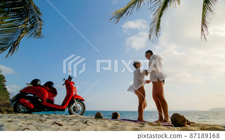 Scooter road trip. Lovely couple on red motorbike in white clothes on sand beach. People walking near the tropical palm trees, sea. Motorcycle rent. 87189168