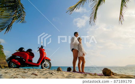 Scooter road trip. Lovely couple on red motorbike in white clothes on sand beach. People walking near the tropical palm trees, sea. Motorcycle rent. 87189170