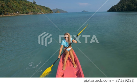 Pretty sportive blonde lady in stylish swimsuit sails on pink plastic canoe along azure ocean bay at exotic resort. Pretty sportive blonde lady in stylish swimsuit sails on pink plastic canoe along azure ocean bay at exotic resort. 87189258
