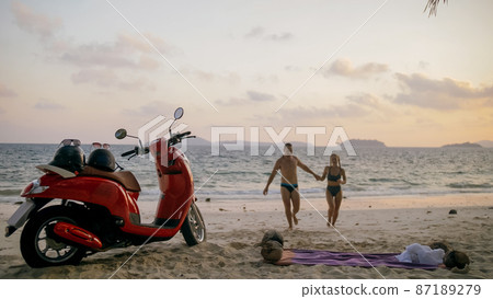 Scooter road trip. Lovely couple on red motorbike in white clothes on sand beach. People walking near the tropical palm trees, sea. Motorcycle rent. Scooter road trip. Lovely couple on red motorbike in white clothes on sand beach. People walking near the tropical palm trees, sea. Motorcycle rent. 87189279