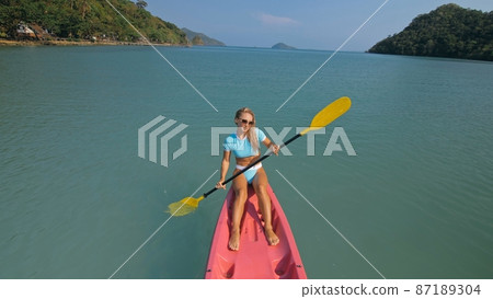 Pretty sportive blonde lady in stylish swimsuit sails on pink plastic canoe along azure ocean bay at exotic resort. 87189304