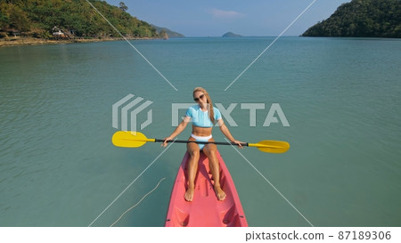 Pretty sportive blonde lady in stylish swimsuit sails on pink plastic canoe along azure ocean bay at exotic resort. Pretty sportive blonde lady in stylish swimsuit sails on pink plastic canoe along azure ocean bay at exotic resort. 87189306