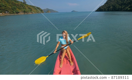 Pretty sportive blonde lady in stylish swimsuit sails on pink plastic canoe along azure ocean bay at exotic resort. 87189311