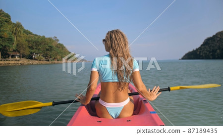 Long curly haired blonde woman in swimsuit rows plastic kayak along azure ocean water at exotic resort close backside view. 87189435