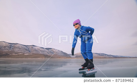 The child train on ice professional speed skating. The girl skates in the winter in sportswear, sport glasses, suit. Outdoor slow motion. 87189462