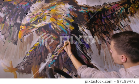 Artist designer draws an eagle on the wall. Craftsman decorator paints picture with acrylic oil color. Painter painter dressed in paint coat. Indoor. 87189472