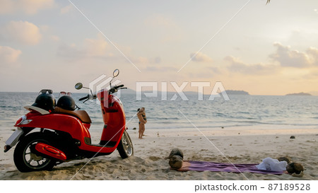 Scooter road trip. Lovely couple on red motorbike in white clothes on sand beach. People walking near the tropical palm trees, sea. Motorcycle rent. 87189528