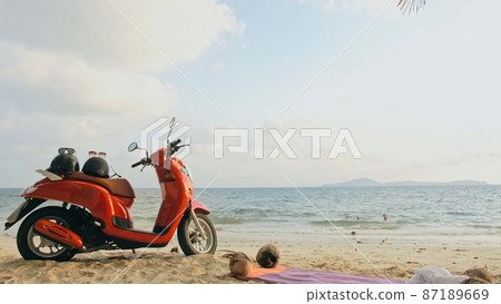 Scooter road trip. Lovely couple on red motorbike in white clothes on sand beach. People walking near the tropical palm trees, sea. Motorcycle rent. 87189669