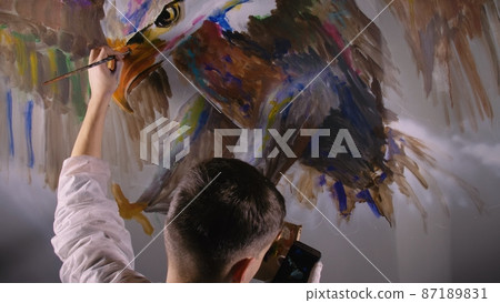 Artist designer draws an eagle on wall. Craftsman decorator paints picture with acrylic oil color looking at sketch in phone. Dark magic cinematic. Artist designer draws an eagle on wall. Craftsman decorator paints picture with acrylic oil color looking at sketch in phone. Dark magic cinematic. 87189831